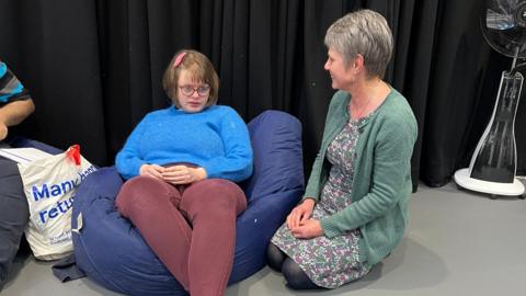 A mother and daughter sat down, daughter sits on a blue beanbag in a blue jumper with brown hair and glasses, mum kneels to her right on the floor with short grey hair, a floral dress and green cardigan