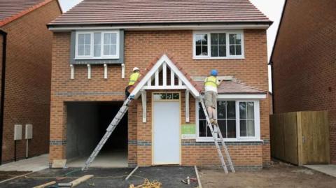 Two men wearing hi-vis vests standing up ladders either side of the doorway of a new build house.