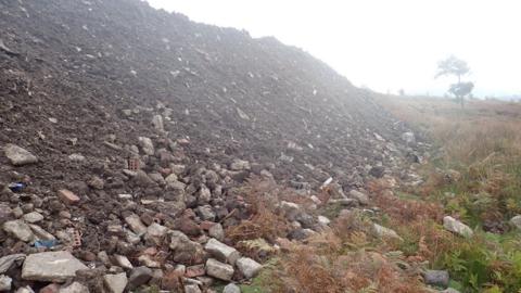 A large pile of rubble and dirt has been dumped in the middle of a grassy field. A tree can be seen in the distance.