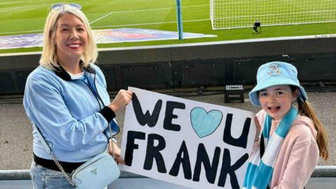 A woman and her daughter stood in the crowd infront of a football pitch holding a sign that says we love you frank. 