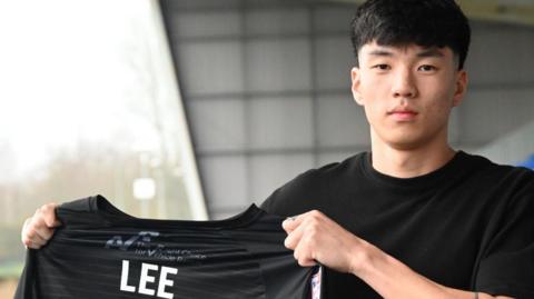 Isaac Lee holds up a black Shrewsbury Town shirt with his surname on the back