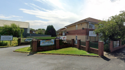 The Gables Care home is a two-storey brick building on a residential road. The main building is slightly hidden behind trees but a sign reads The Gables Care 鶹Լ.