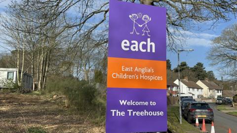 A large roadside sign for East Anglia's Children's Hospices, also known as EACH. The sign welcomes visitors to a facility called The Treehouse. It stands on a grassy verge beside a road.