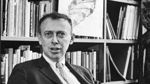 Dr James D Watson sitting at his desk in front of a drawing of the double helix structure of DNA, early 1960s