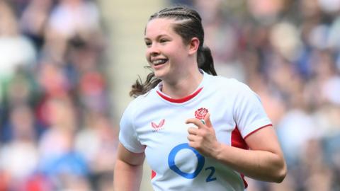 Demelza Short holds her left arm bent across her body and smiles while jogging on the pitch wearing an England shirt