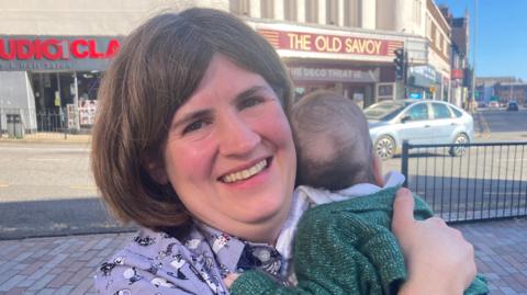 Zoe Smith holding her baby against her shoulder in Northampton town centre. She has shoulder-length brown hair, a lilac top that has illustrations of cats on it, and is smiling at the camera. Her baby is facing away from the camera and is wearing a dark green cardigan.