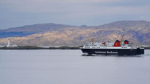 The MV Isle of Lewis is a large black and white ferry with two red funnels either side of each other and yellow and red logos. The ship is sailing in calm waters past low rugged hills.