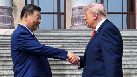 Xi JInping (left) wearing a navy blue suit, smiles as he stretches his hand towards Donald Trump, who is also in a navy blue suit