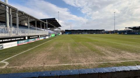 A view of Barrow AFC's home ground at SO legal stadium. The view is taken from near one of the corner stands and shows much of the pitch. The grass near the side-line has been warn and is patchy. A stand with rows of blue seats can be seen to the left.