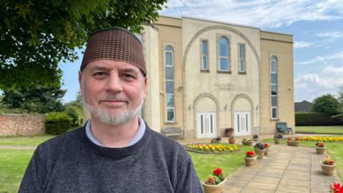 A man, smiling, in front of a mosque; he has a short, white beard and is wearing a taqiyah cap.