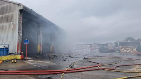 A forecourt with fire hoses and some rubble across it. Smoke pours out of a large warehouse.