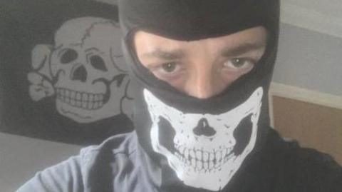 Alfie Coleman wearing a black balaclava with a skull on it. Only his eyes can be seen. Behind him a black flag with a white skull and crossbones is pinned to a wall.