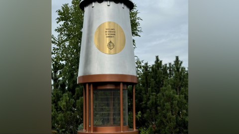 A tall, silver and orange-looking lamp stands on a traffic island. It has a gold circle on it which says the lamps are dedicated to the mining community. Light comes from the middle section.