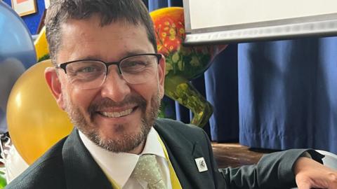Craig Jensen has short brown hair and is wearing black framed glasses, a white shirt, black suit jacket and green tie. He is holding a silver metal time capsule to the right.