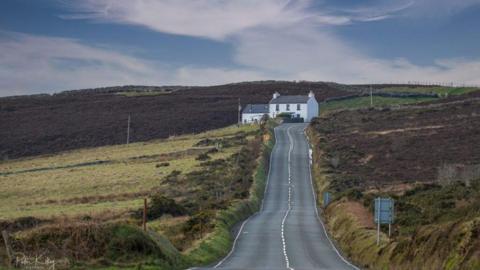 A long winding road with an embankment on the right, and green hills on the left, a white cottage is on the right of the road at the top. 