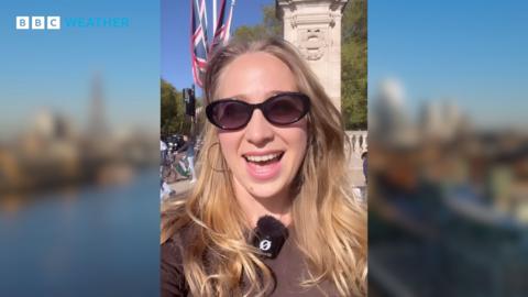 Woman wearing sunglasses standing beneath bright blue sky with Union Flag behind