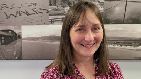 Dr Rowena Christmas has brown shoulder length hair and a partial fringe. She has blue eyes and is smiling at the camera. She is wearing a patterned shirt with different shades of pink and is stood in front of some black and white beach photos