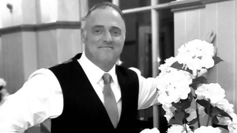 Ray McDonald, a man in his 60s, is wearing a white shirt, a black waistcoat and tie whose colour is unclear as the image is in black and white. He is standing in a room that could be a hotel or function room, with a large floor-to-ceiling window behind him, next to an arrangement of apparently white flowers. He is smiling.