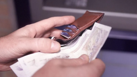 Two hands holding brown leather wallet withdrwaing banknotes. The card slots are full of cards.