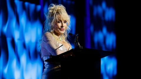 Dolly Parton in a gold dress with blond poufy hair stands at podium against a blue background