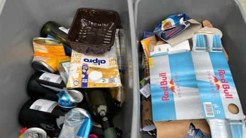 Two grey boxes containing recyclable waste. The one on the left contains plastic, cans, glass bottles and cartons. The one on the right contains paper and cardboard.