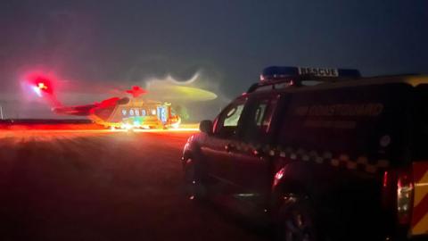 A Coastguard Rescue helicopter with its lights flashing pictured stationary next to an emergency vehicle.