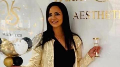 A person stands in a bright, modern interior beside a white wall featuring a gold logo and the text “AESTHETICS.” The person is wearing a black outfit with a gold belt and a gold sequined jacket, holding a large transparent balloon