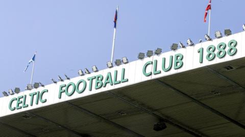 The top of the Celtic Park stadium, with flags on the roof and a green sign which reads Celtic Football Club 1888