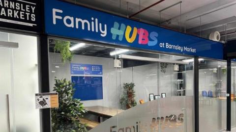 A unit in Barnsley Market building with a blue sign and multi-coloured lettering spelling out the words Family Hubs. A plate glass window reveals tables and seating inside.