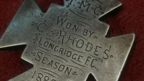 A silver medal in a four sided cross shape engraved with the letters TMC and then "Won by C. Rhodes, Longridge FC, Season 1885 -6".