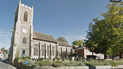 A medieval church in a town centre, with gravestones in the foreground, a tree to the right, and a set of traffic lights and a road to the left 