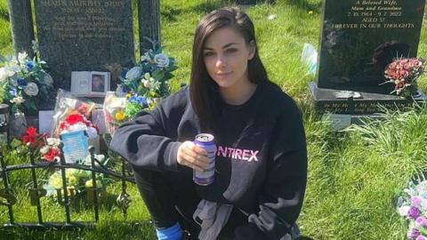 Lauren kneels beside a decorated graveside in an all black outfit. She holds a can.