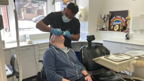 A man in a blue hoodie has his mouth open and is being examined by a dentist, wearing a mask and gloves and a black top. On the wall of the examination room is a sign with 'save the NHS' and a number of trophies and boxing belt.
