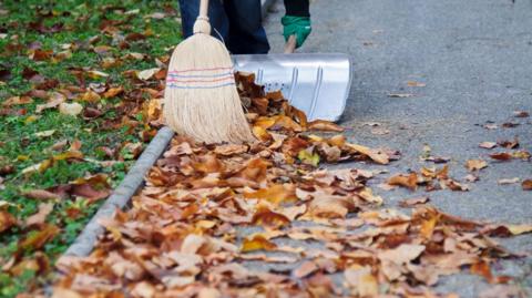 Orange leaves on tarmac beside a grass verge are being swept up by a straw brush, into a metal shovel, by someone wearing blue overalls and green gardening gloves.