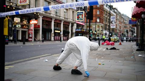 A street closed by police cordons on both ends. A forensic officer in while overalls is picking up an item from the pavement. 