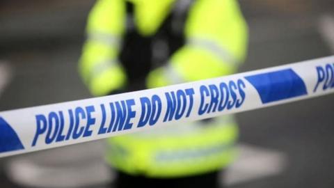 Police officer stands behind police tape reading police in blue on white background