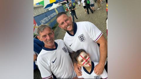 Two men (Gary and Ben) and a boy wearing white England shirts. The boy (Ronnie) has the England flag painted on his face