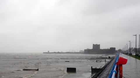 A stormy seaside promenade with rough waves, grey sky, and a distant castle in the rain.
