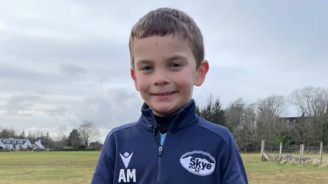 Archie na sheasamh air pàirce agus gàire mhòr air aodann. 