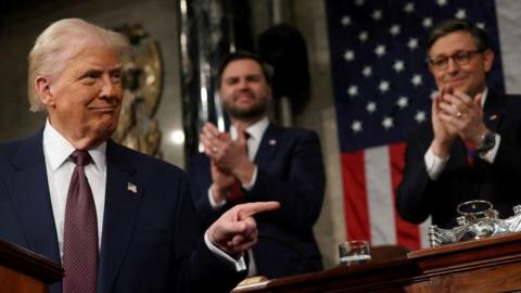 President Trump wearing a suit and tie, smiles and points his finger, with JD Vance behind him
