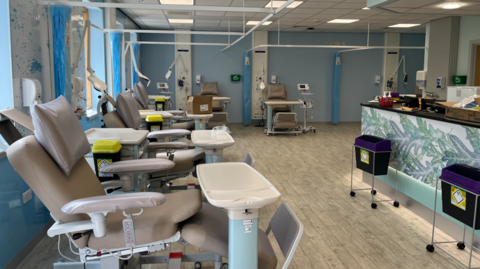 A hospital room with a desk on the right hand side, and five grey reclining chairs along the left hand side and back of the room, each with a medical table in front of it.