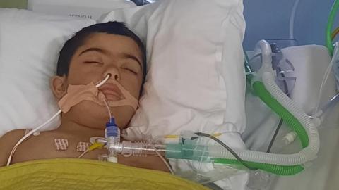 A young boy with dark hair is lying unconscious in a hospital bed with tubes coming out of his mouth and nose. A hospital monitor is at the head of his bed. A brightly coloured family blanket is on the bed cover. 