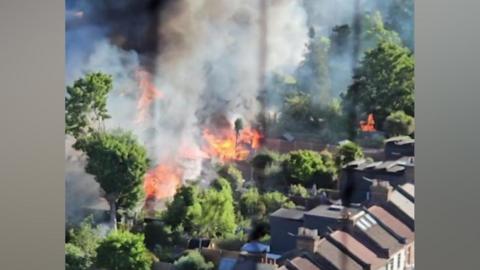 A high-angle, slightly blurred photo showing a serious fire burning intensely amongst green trees and back gardens behind a row of terraced houses, with thick columns of dark smoke filling the air.