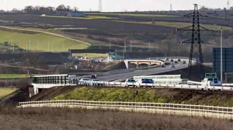A dual carriageway road snakes across a rural landscape of green and brown fields, dotted with electricity pylons, under a grey sky. Several bridges can be seen.