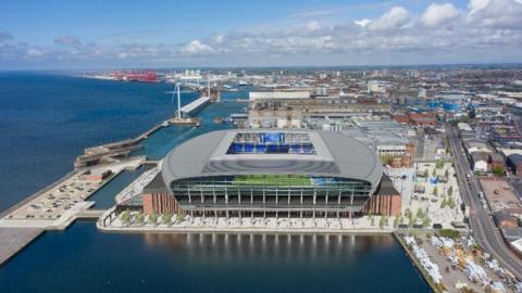 A large stadium sits on a dock with the River Mersey to the left and the outer city of Liverpool to the right