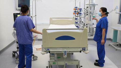 An empty bed in an intensive care unit in a hospital. Two people in blue medical scrubs are standing either side of it. There is medical equipment around the bed. The room is white.