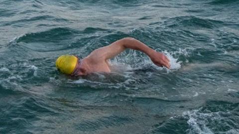 John Bauer swimming in the sea wearing a yellow swimming cap and clear goggles.