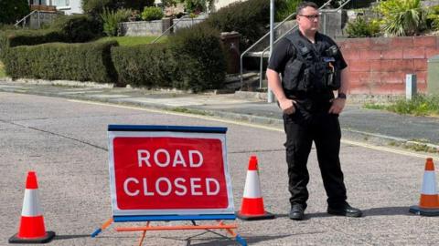 A police officer standing to the right of a red 'ROAD CLOSED' sign in the middle of a road. The officer is wearing all black and has his hands in his pockets. There are orange and white traffic cones either side of the sign.