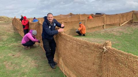 A group of people seen building a fence on grassland. One person in a dark blue coat is seen looking at the camera and smiling. The sky is grey and everyone is wearing winter coats.