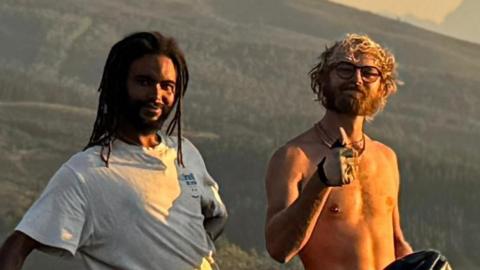Two young men stand in front of a grassy hilly area and smile at the camera. One wears a white T-shirt and has black dreadlocks. The other has curly blonde hair, is shirtless, has glasses on and raises his thumb.
